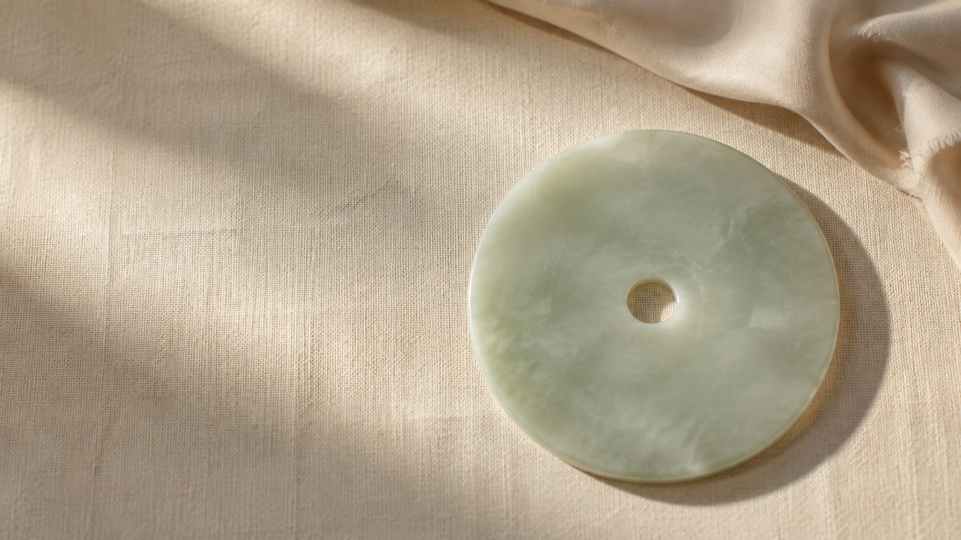 An illustration of a pale green jade ring resting on linen beside a fold of raw silk, lit by soft daylight.