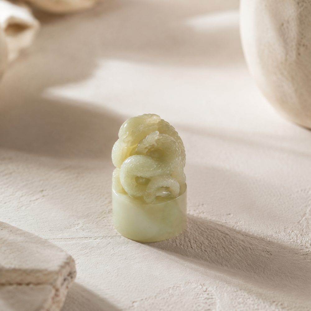 An illustration of the handle of a jade seal, carved as a knot of coiled dragons, in pale green nephrite against a dark ground. The stamping face is not shown.