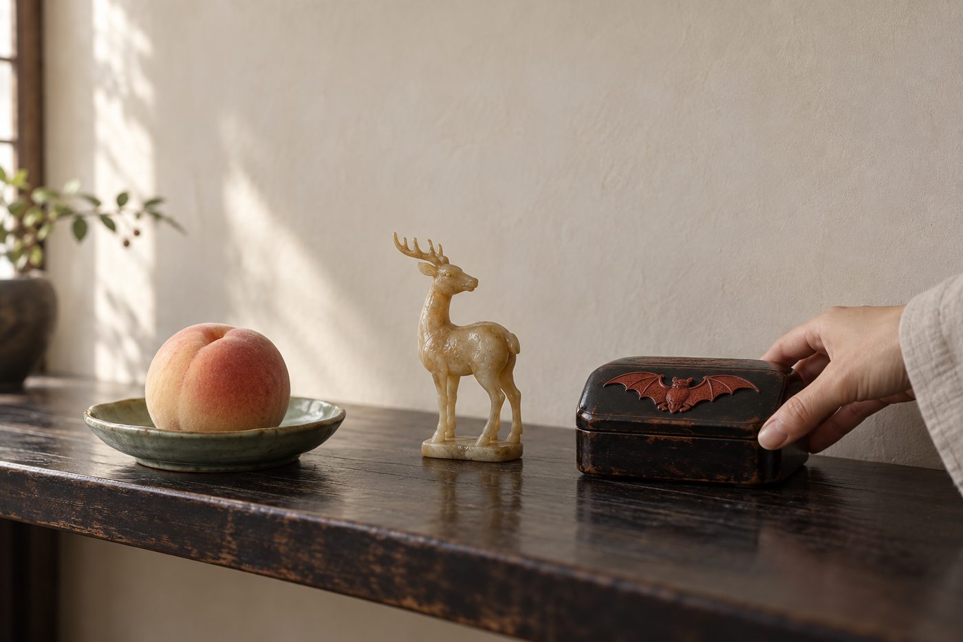 A peach on a celadon dish, a small carved deer, and a black lacquer box with a red bat on its lid, arranged on a shelf as a hand straightens the box.