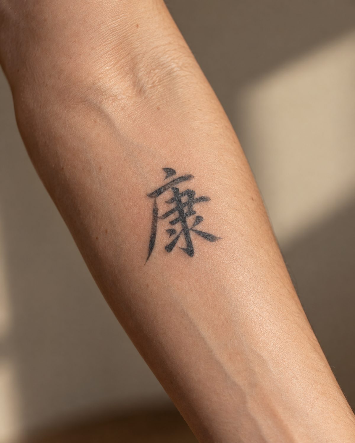 The Chinese character for good health, 康 (kāng), as a fine brush-style tattoo on an inner forearm.